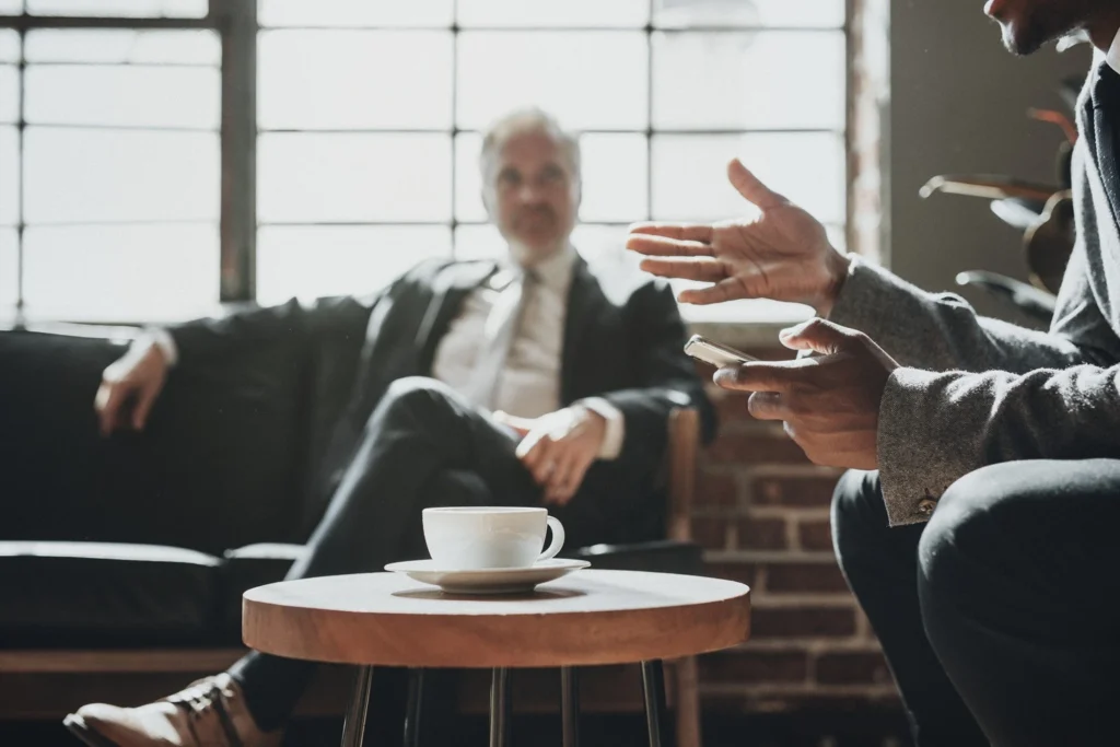 Two professionals having a conversation in a modern office setting, with a cup of coffee on a table in the foreground.