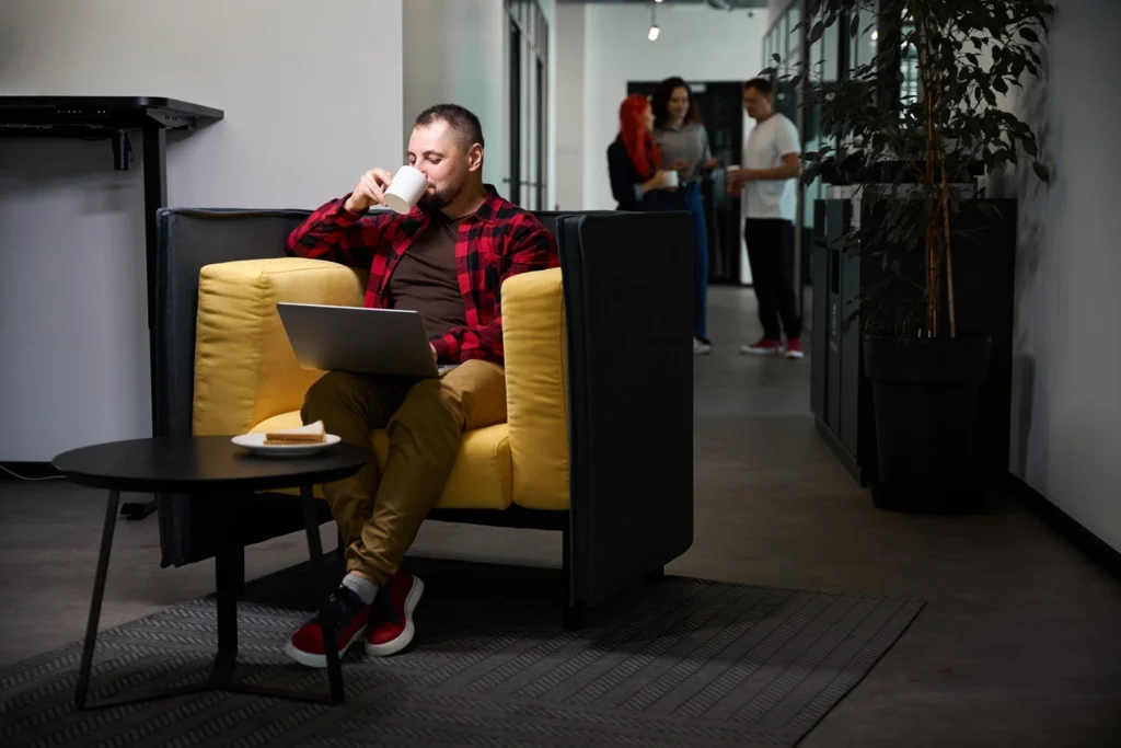 Engineer in an Irish tech office drinks coffee while working on a laptop in a lounge area, colleagues chatting nearby; on-site coffee helps focus and connection.
